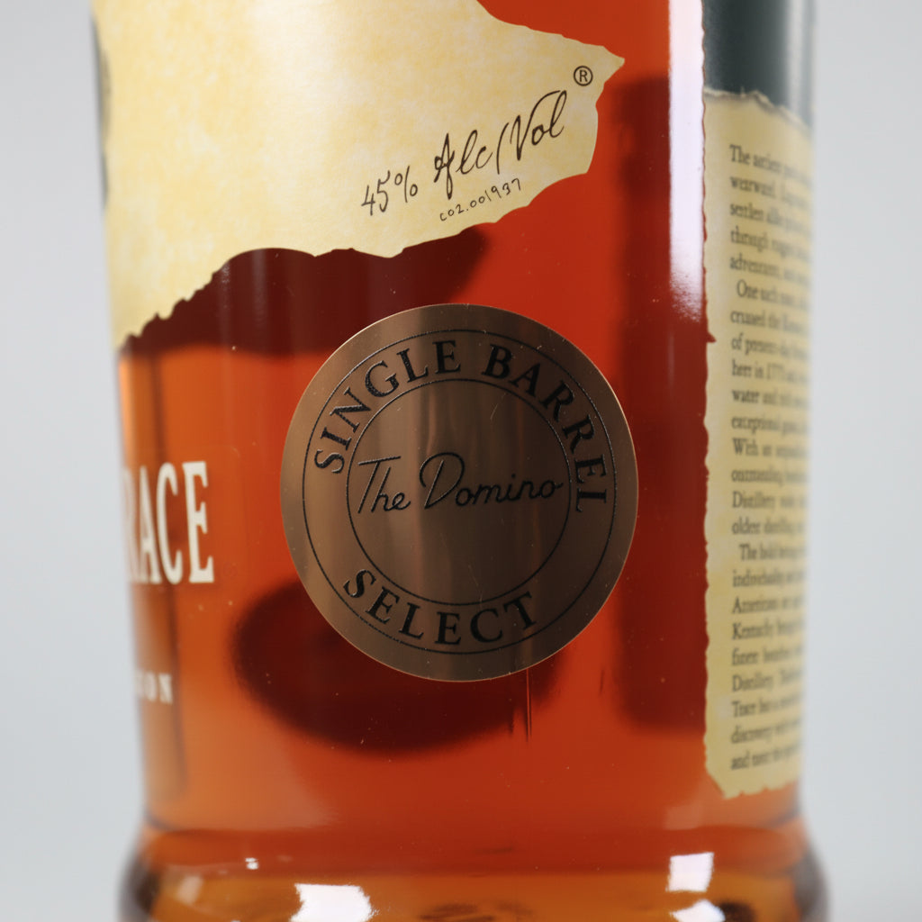 Close-up of a whiskey bottle showing amber liquid, a gold label with 'Single Barrel Select' and 'The Dominio', and part of a cream paper label on a white background.