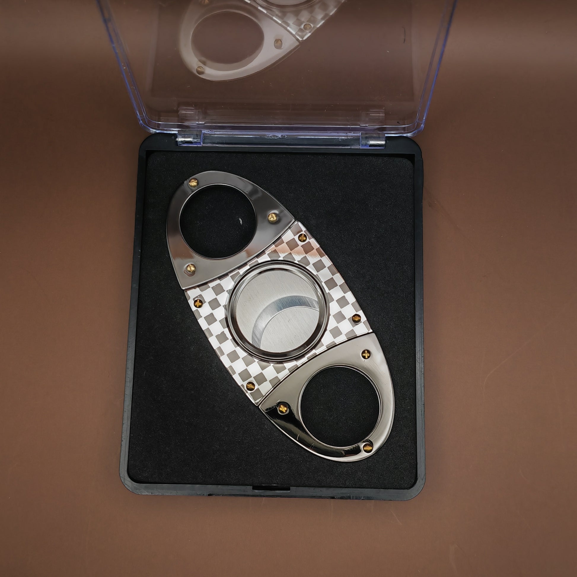 Cigar cutter with checkered design on a black mat inside an open clear case on a brown background