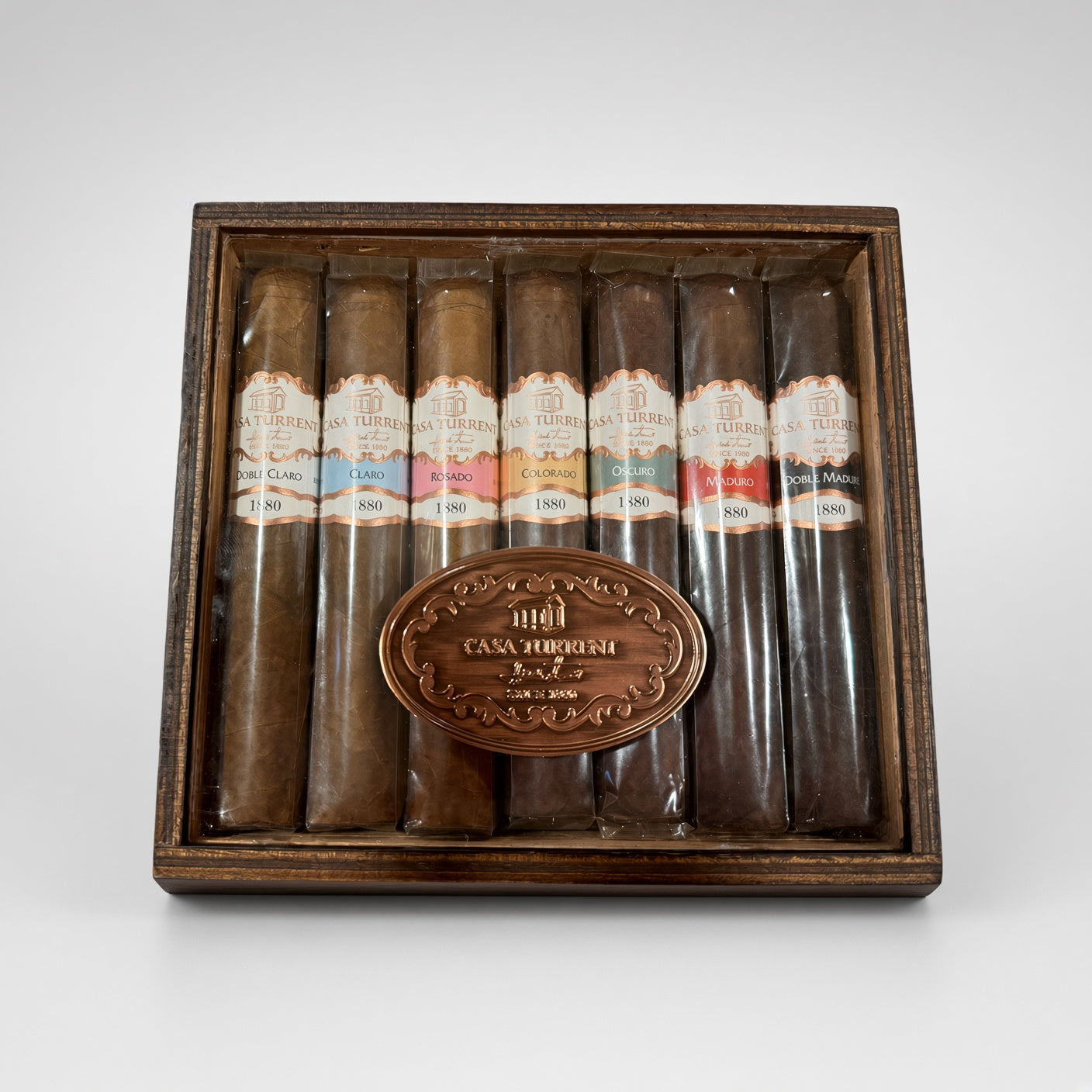 A premium cigar gift set in a dark wood box, featuring eight cigars of varying shades from light brown to very dark brown, each with a colorful label, and a central oval bronze plaque engraved with 'CASA TOROUSH'.