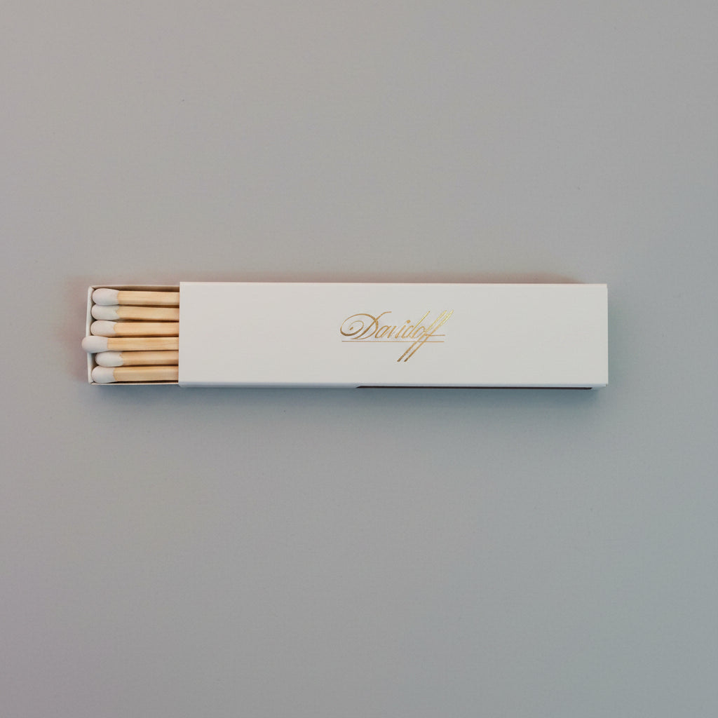A pack of wooden matches in a white rectangular box with gold foil script, placed on a textured gray surface.