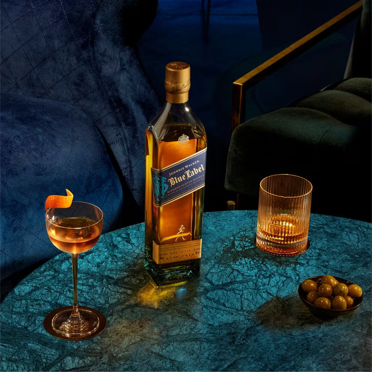 A bottle of Johnnie Walker Blue Label whisky stands on a round teal table in a dimly lit lounge setting, with a filled coupe glass, an empty textured tumbler, and a small bowl of olives nearby.