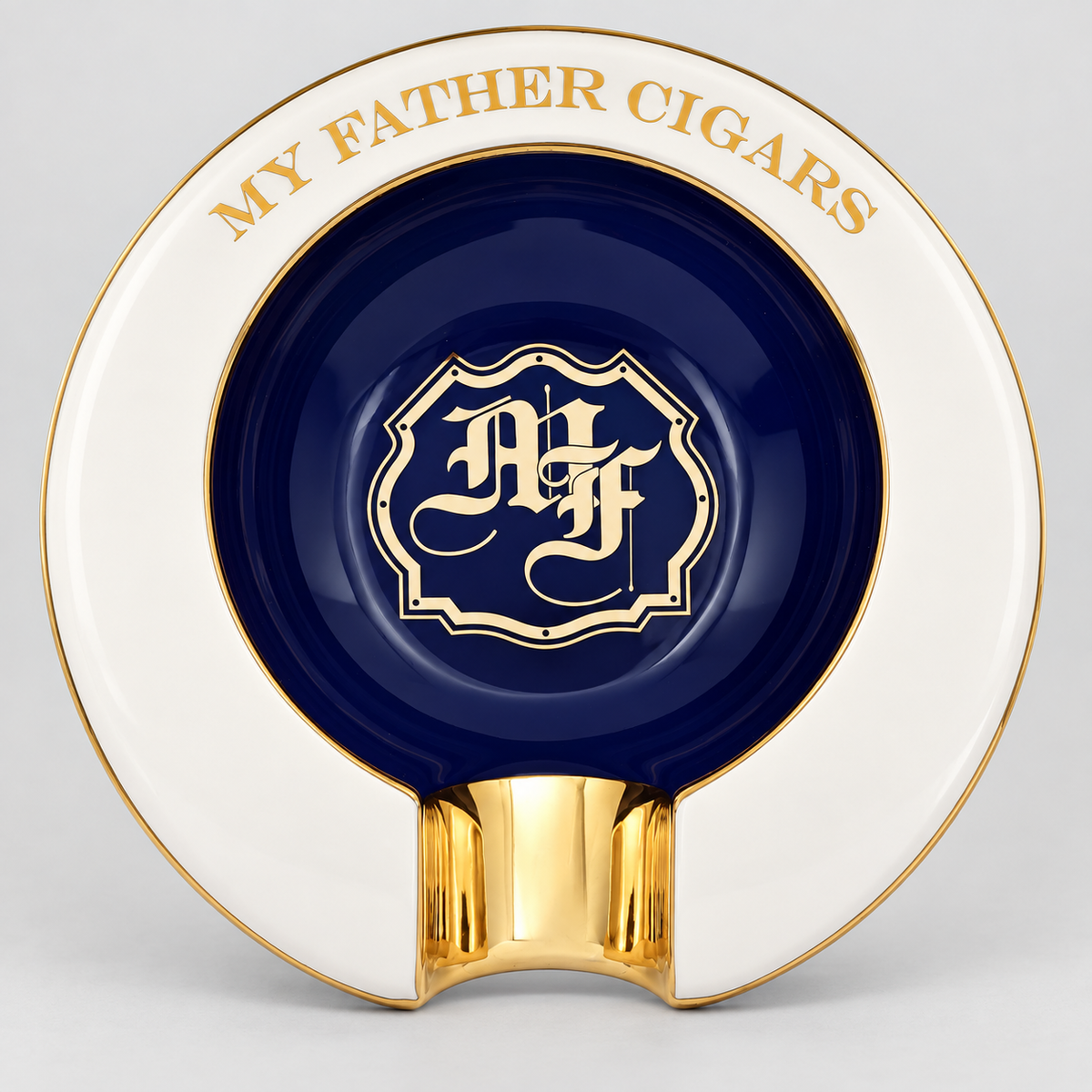 A round ceramic cigar ashtray with a white outer rim, a dark navy blue center, and polished gold detailing. The rim is inscribed with the text 'MY FATHER CIGARS' in gold, and the center features a gold crest containing the initials 'AF'.
