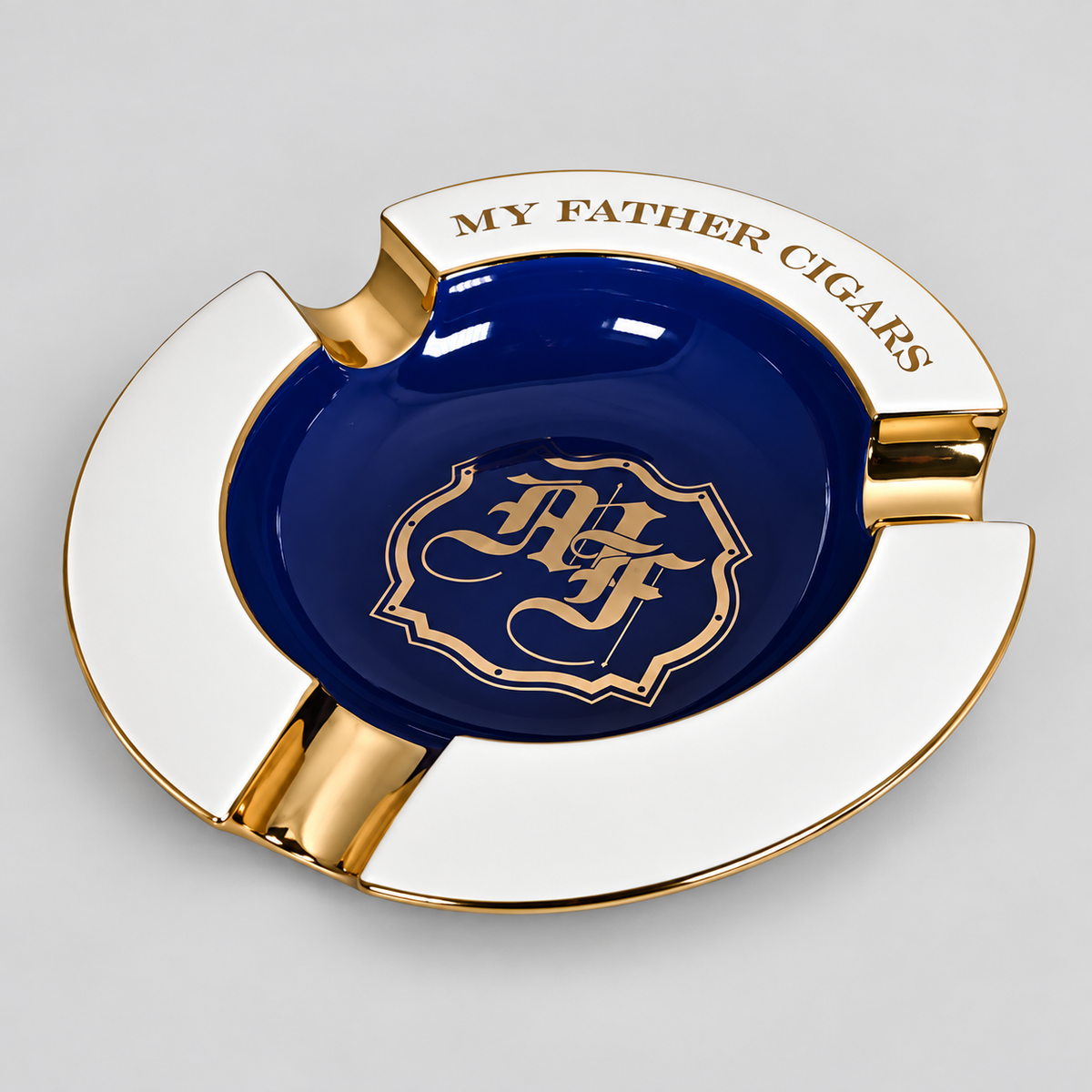 A round ceramic cigar ashtray with a glossy white outer rim, a deep navy blue center, and polished gold accents. The top edge features the text 'MY FATHER CIGARS' in gold, and the center is decorated with a gold crest containing the initials 'AF'.