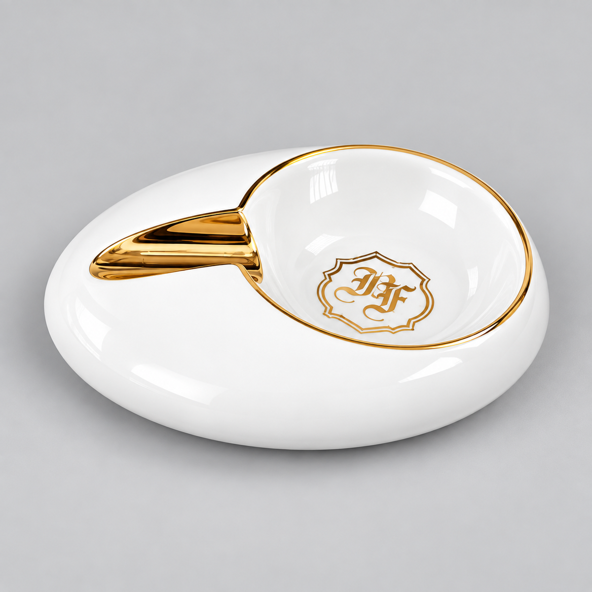 A white ceramic ashtray with a gold-trimmed handle and a gold crest emblem inside the bowl, shown on a neutral gray background.