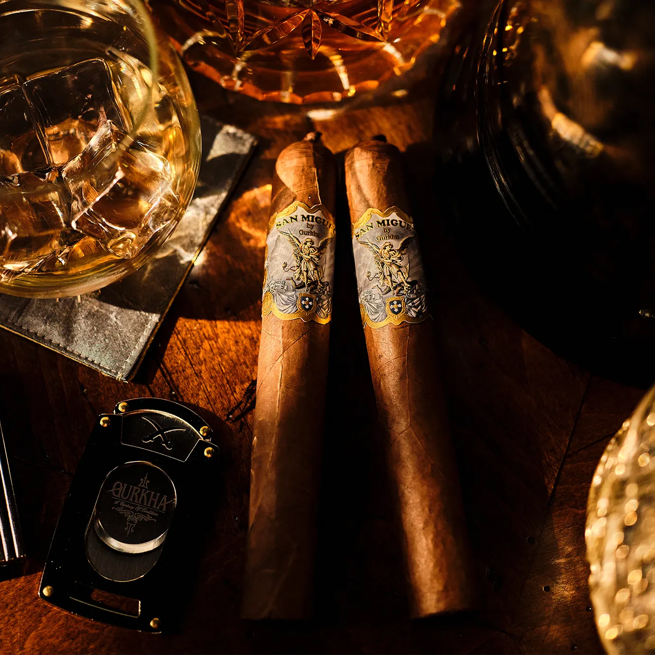 Two cigars with a lighter and a glass of whiskey on a wooden surface.