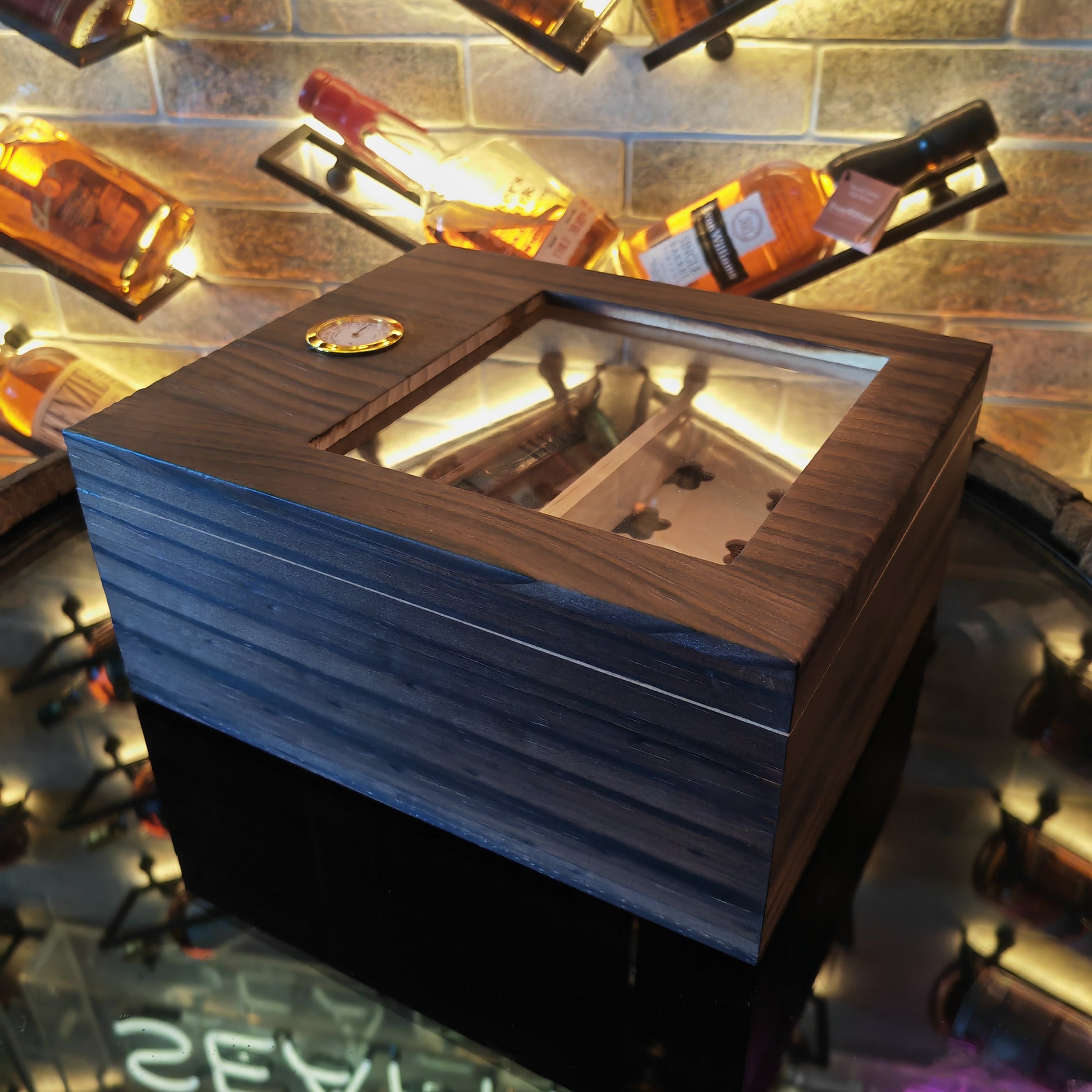 Wooden cigar box with glass top on a reflective surface with bottles in the background