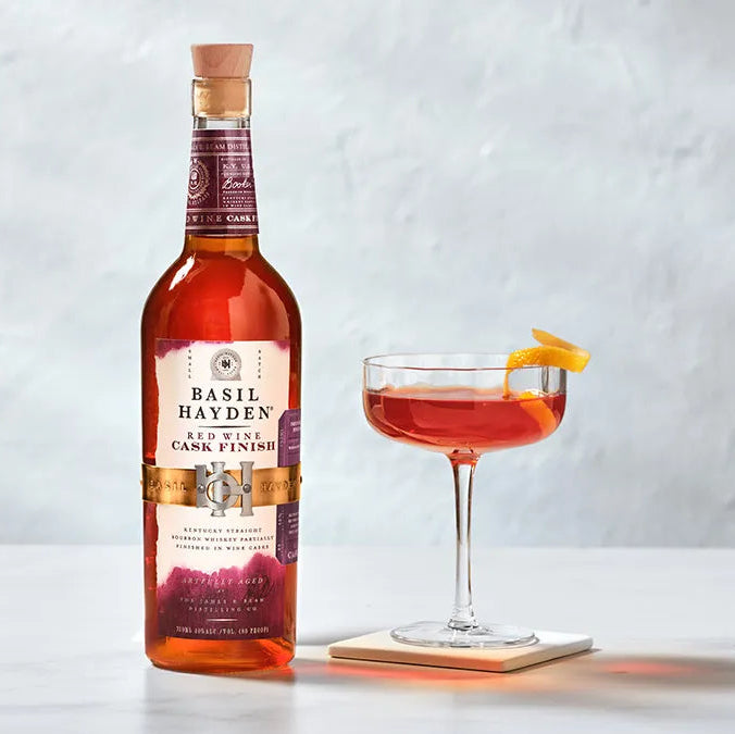 Bottle of Basil Hayden whiskey and a cocktail with an orange twist on a light gray background