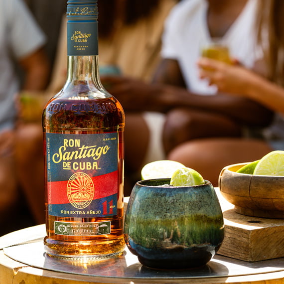 Bottle of Ron Santiago de Cuba rum with a glass and lime on a table, people in the background.