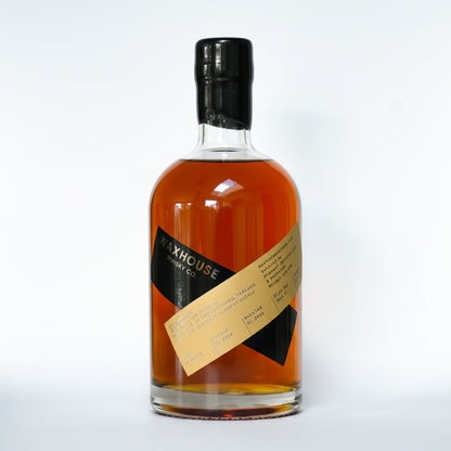 A bottle of Waxhouse Whiskey Co. AMERICAN RYE whisky, showing the amber liquid through a clear glass bottle with a black cap and a diagonal tan and black label.