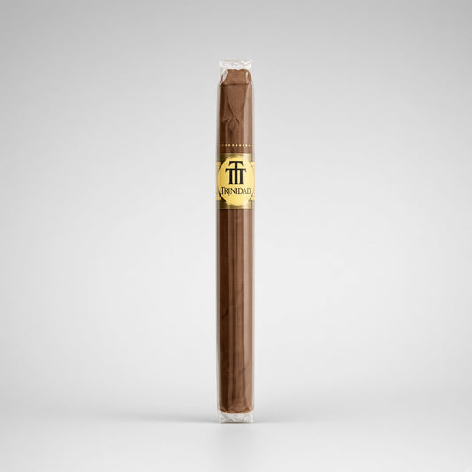 A single Trinidad cigar in its clear plastic wrapper, showing a dark brown wrapper body with a gold and black logo band, set against a light gray studio background.