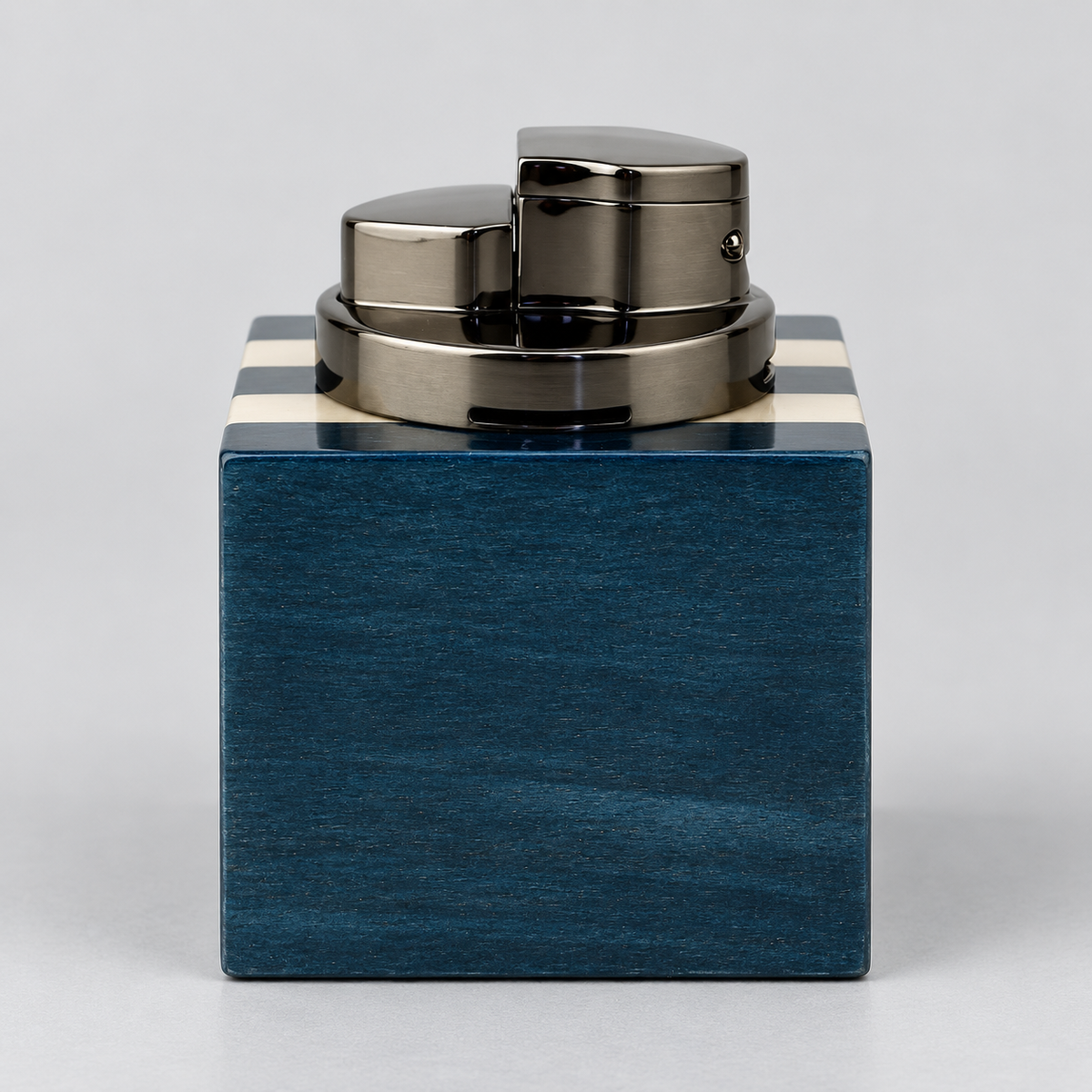 A luxury lighter with a polished gunmetal gray metallic body and a square-shaped dark blue wood base featuring cream-colored stripes on the top edges, set against a neutral light gray background.