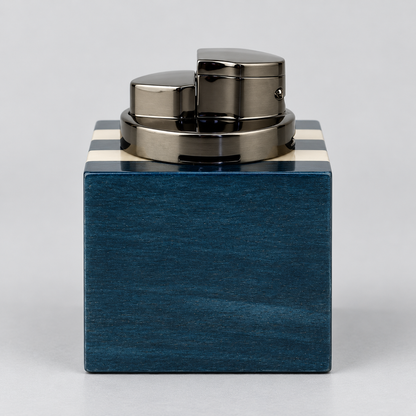 A luxury lighter with a polished gunmetal gray metallic body and a square-shaped dark blue wood base featuring cream-colored stripes on the top edges, set against a neutral light gray background.