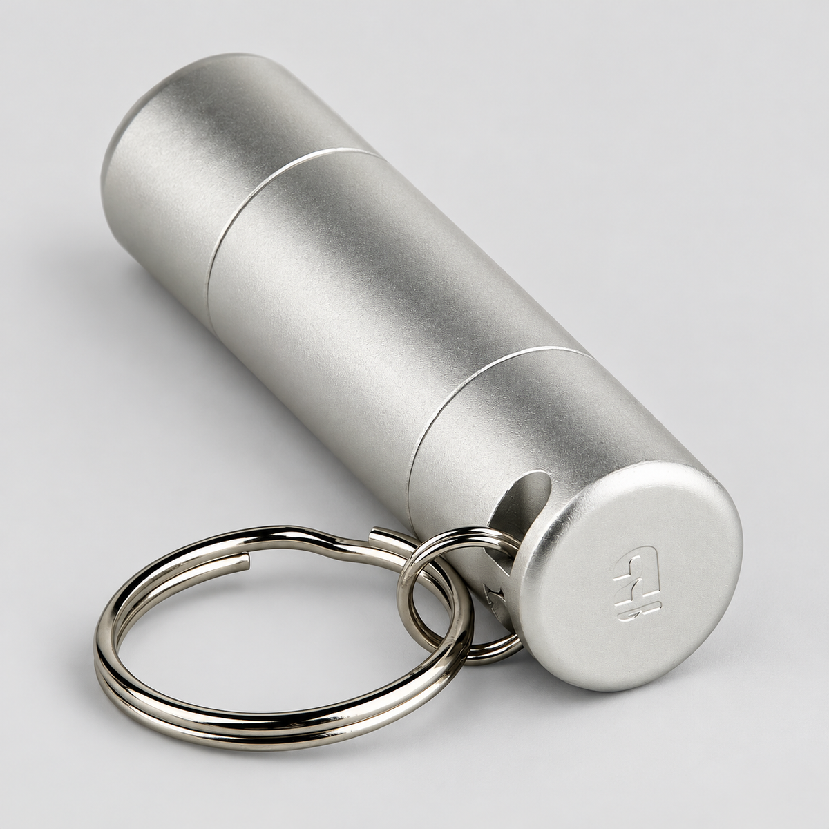 A silver aluminum cylindrical keychain container with a matching split key ring, shown on a light gray background.
