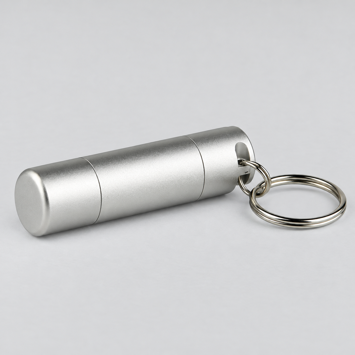 A silver aluminum cylindrical keychain container with a matching split key ring, shown on a neutral light gray background.
