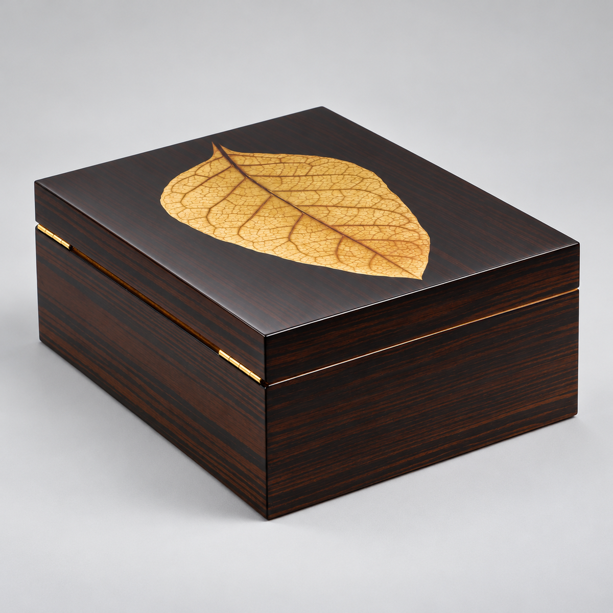 A square dark wood presentation box with a glossy finish, featuring an inlaid golden-brown leaf on the lid and a gold-tone metal clasp on the front edge, set against a neutral gray background.