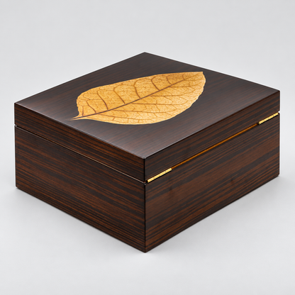 A square dark brown wooden box with a glossy grain, featuring a decorative golden-yellow leaf inlay on the lid and a thin gold metal accent along the front edge, set against a neutral light gray background.