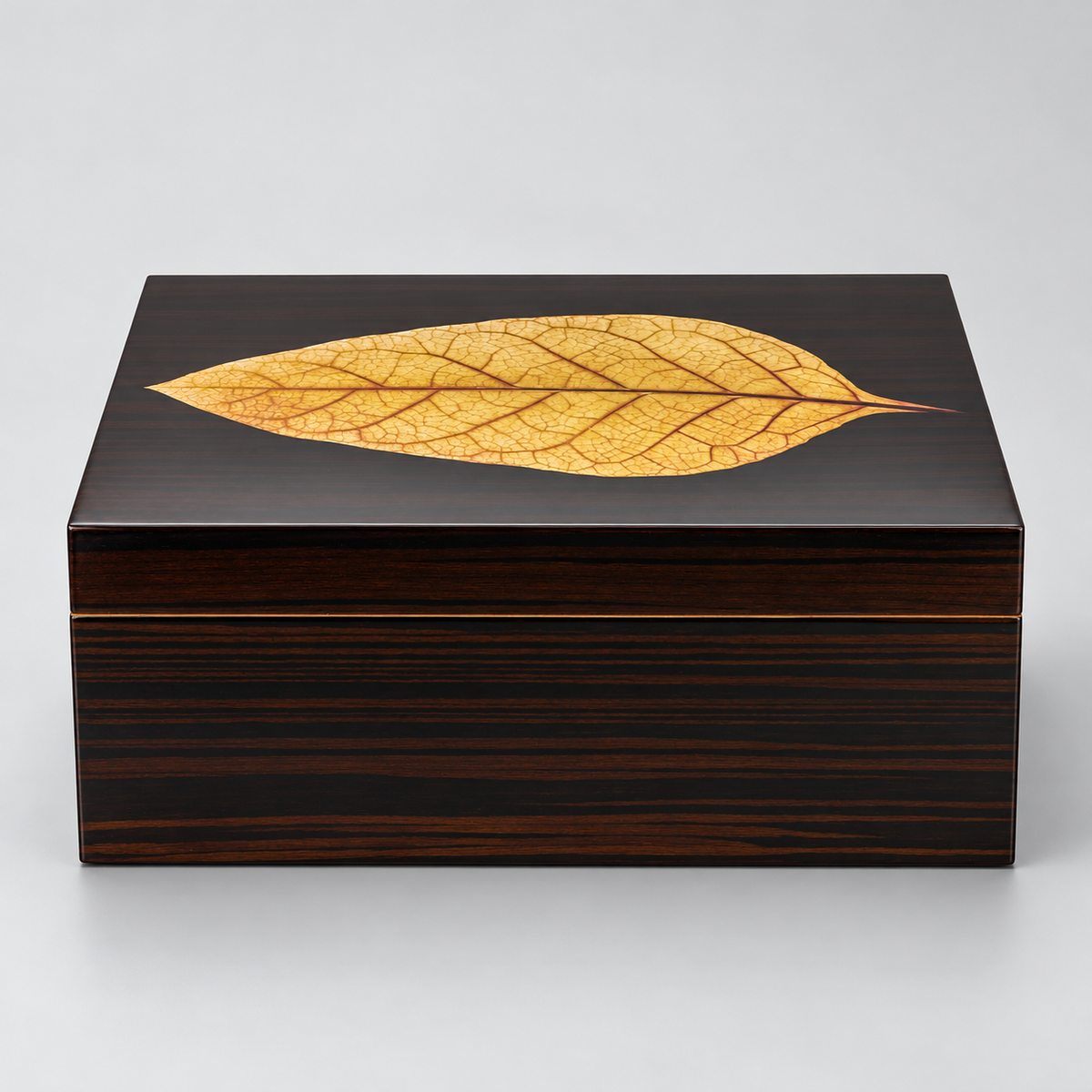 A rectangular dark wood presentation box with a glossy black top featuring a large, detailed golden-yellow leaf inlay, set against a neutral light gray background.