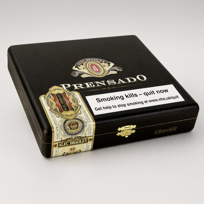 A black cigar humidor box by Prensado, featuring gold and white decorative branding, a gold clasp, and a prominent white smoking-health warning label on the front, photographed on a light gray background.