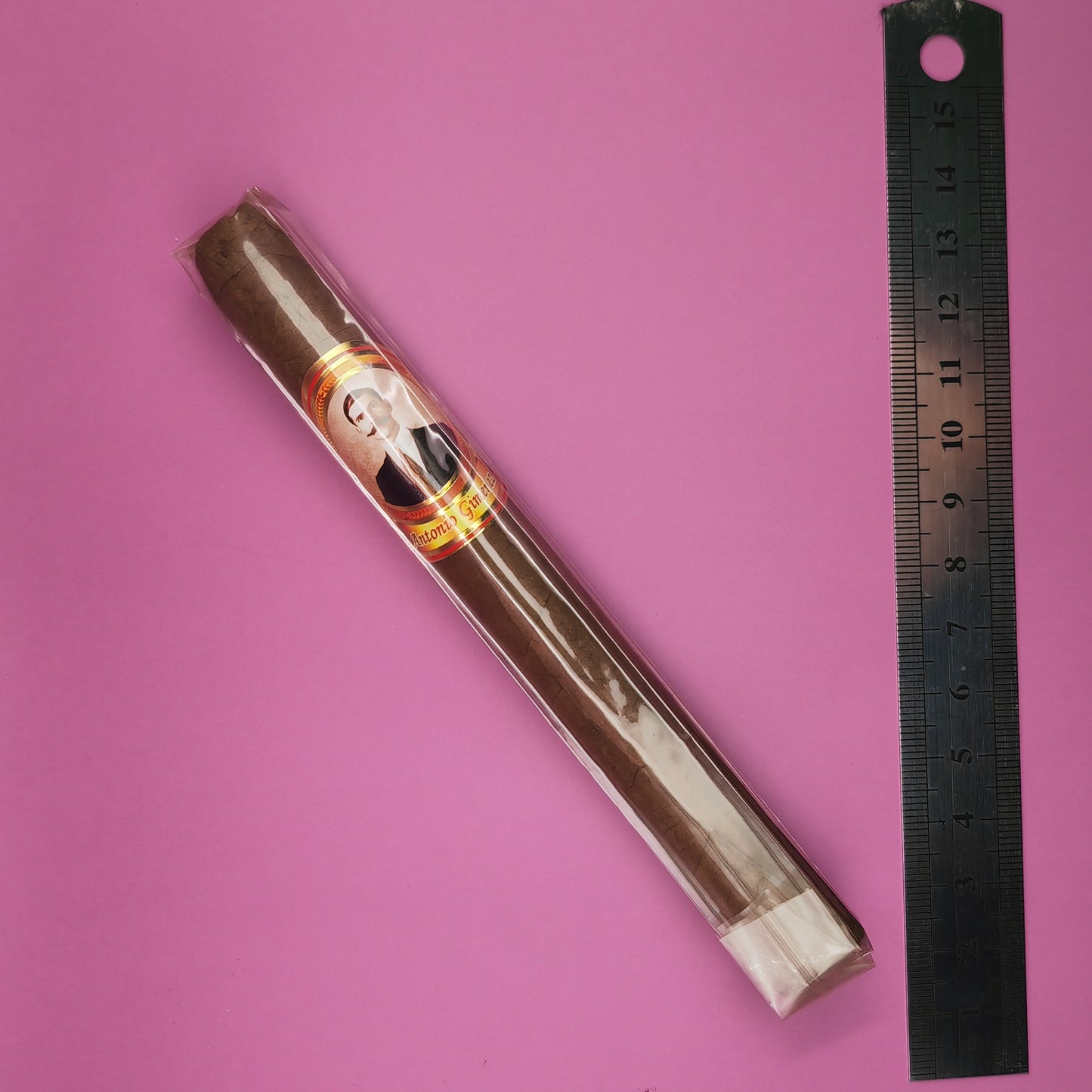 Cigar in a clear plastic tube with a brand label next to a ruler on a pink background