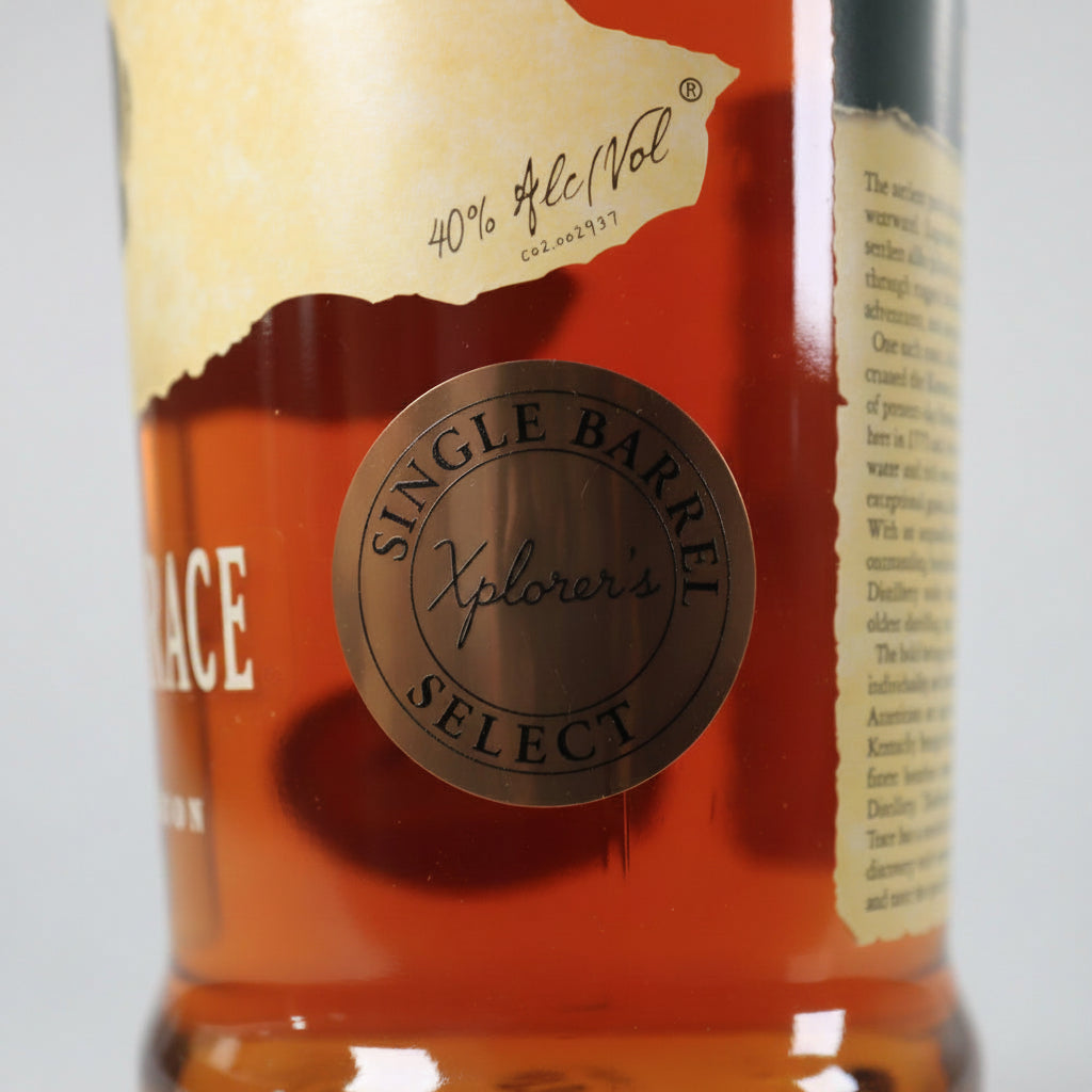 Close-up of a whisky bottle label showing a bronze circular seal that reads 'Single Barrel Xplorer's' over amber liquid, with part of a beige label and white text visible.