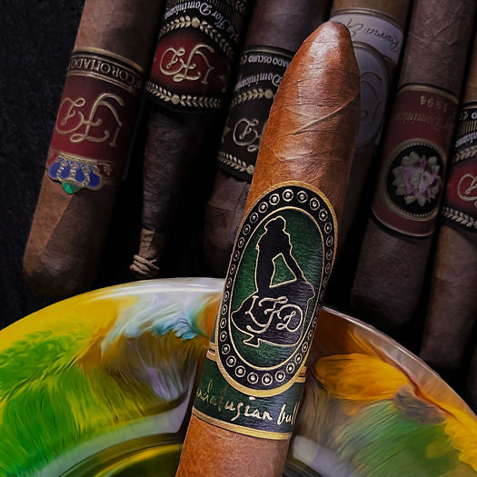 A single brown cigar with a green and black band featuring a silhouette of a woman and the text 'Cigaran Bolivar', displayed in front of a colorful, feathered ashtray with other cigars in the background.