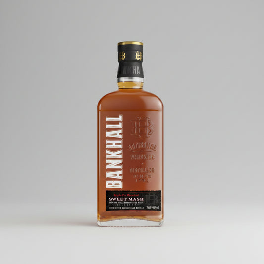 A bottle of Bankhall Sweet Mash Blackpool american style Whiskey against a neutral gray background. The clear glass bottle showcases the rich amber-brown liquid inside, with a black and gold neck seal and a black label featuring vertical white text.