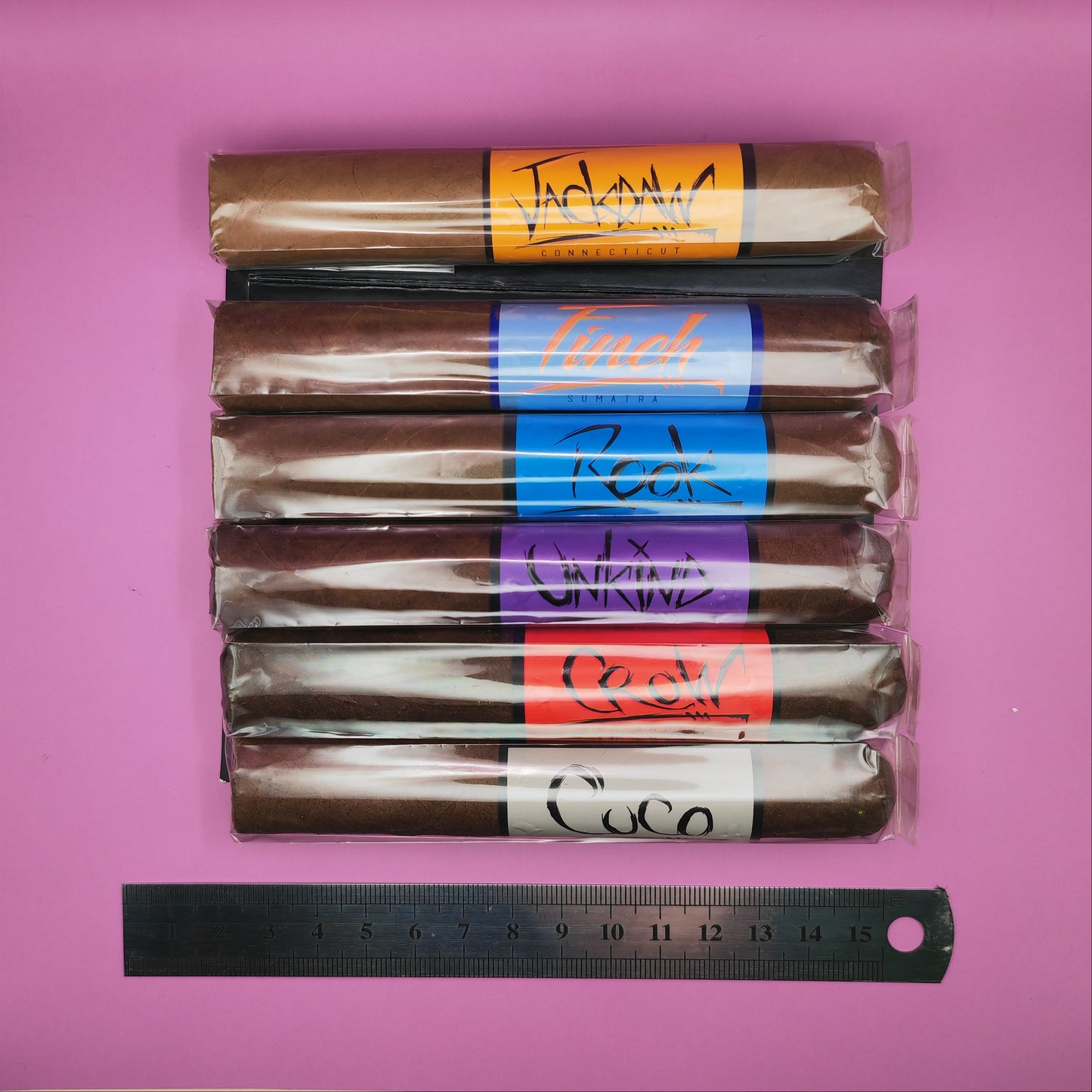 Packaged cigars with visible brand labels on a pink background, accompanied by a ruler for scale.