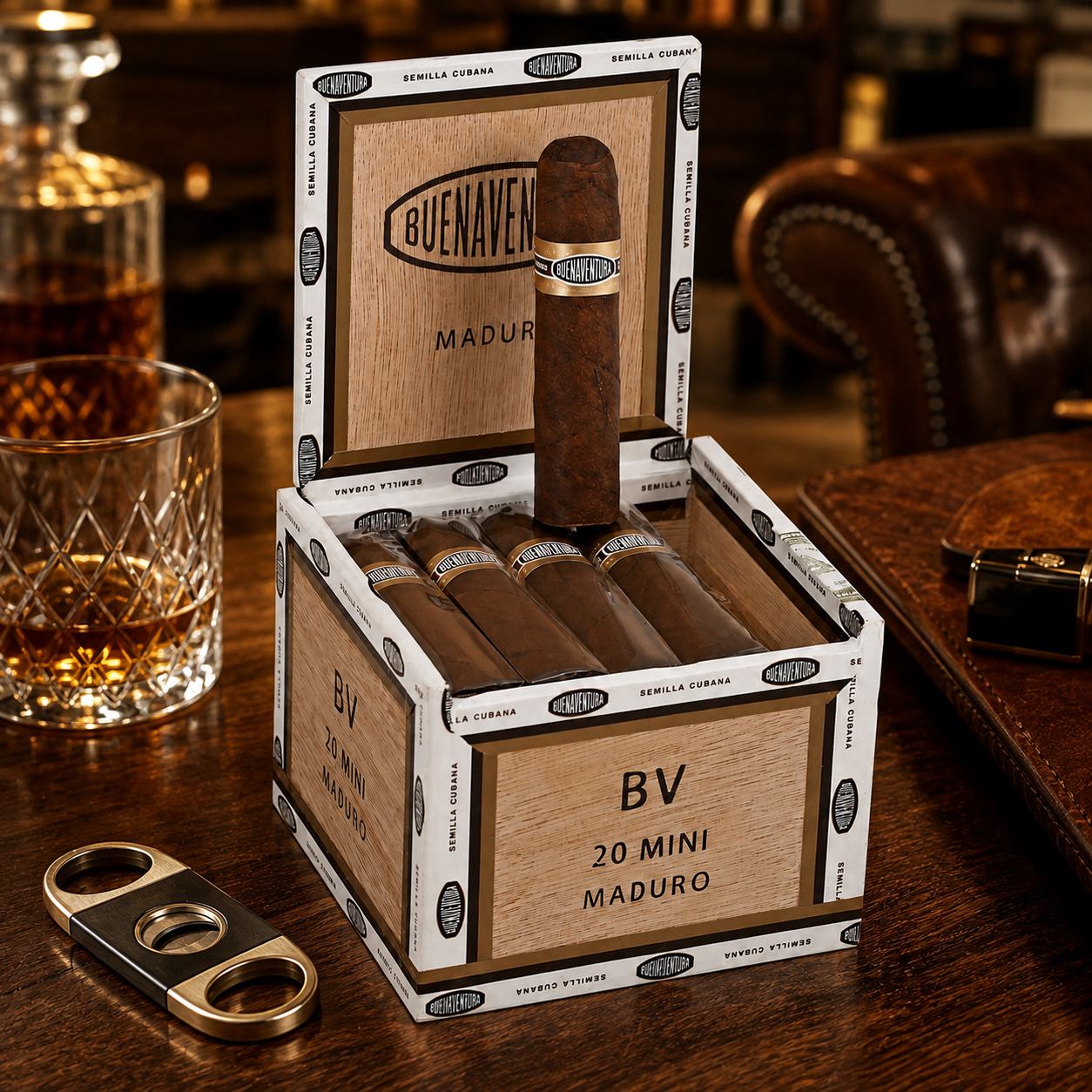A box of Buena Ventana 20 Mini Maduro cigars on a dark wooden table, with a single cigar standing upright in the open box. A cigar cutter lies nearby, and a glass of amber liquor and leather accessories are visible in a bar-like setting.