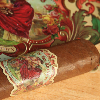 A close-up of a brown cigar with a colorful vintage-style label reading 'Sun Grown' and 'Cigarillo', resting on a light wood surface next to a matching branded wrapper roll.