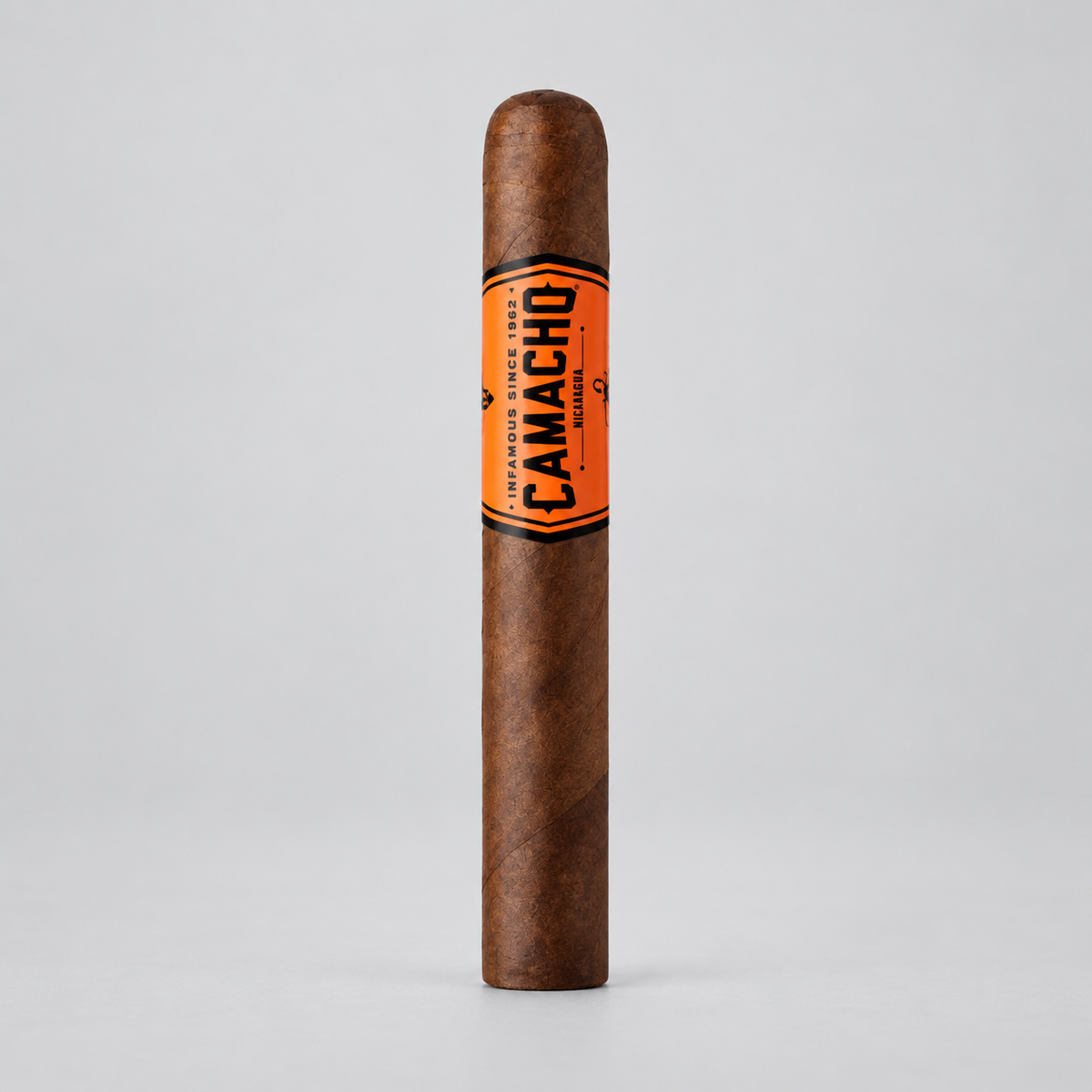 A single Camacho cigar standing upright on a light gray background, featuring a brown tobacco body with a bright orange band displaying the black Camacho brand name.