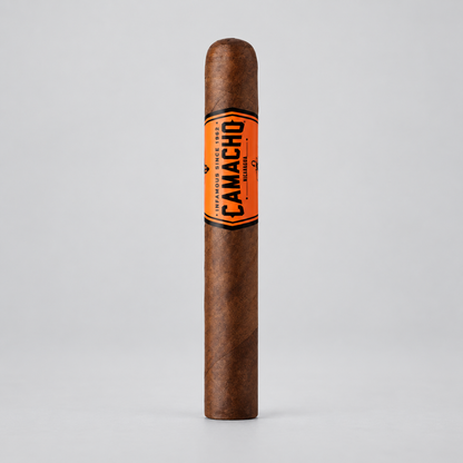 A single Camacho cigar standing upright on a light gray background, featuring a brown tobacco body with a bright orange band displaying the black Camacho brand name.