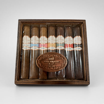 A premium cigar gift set in a dark wood box, featuring eight cigars of varying shades from light brown to very dark brown, each with a colorful label, and a central oval bronze plaque engraved with 'CASA TOROUSH'.