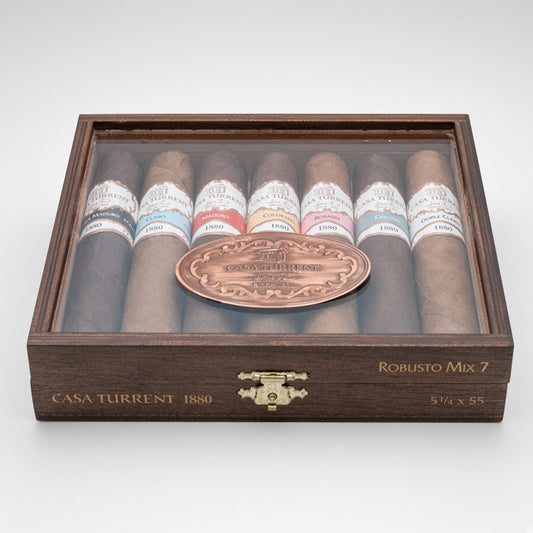 A premium cigar gift set in a dark wood display box with a clear lid, showcasing eight cigars with various colored bands and a central copper-toned oval medallion. The box is branded 'Casa Turrent 1880' and labeled 'Robusto Mix 7'.