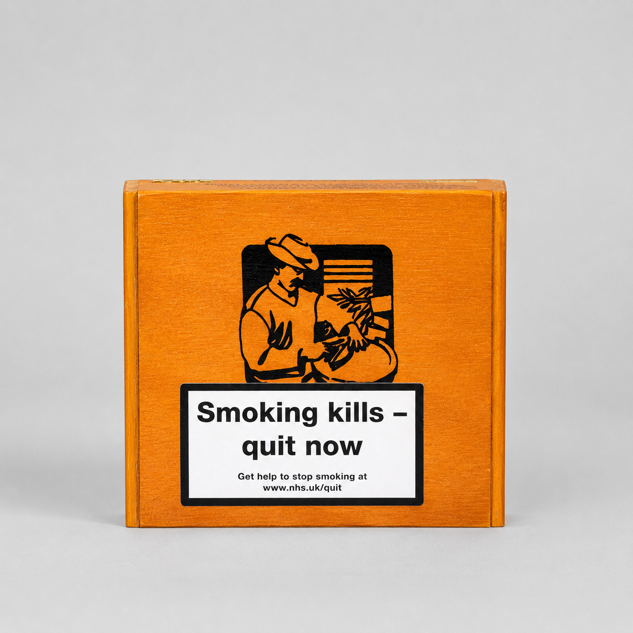 A square wooden box with a warm orange-brown finish, featuring a black graphic of a person smoking a cigarette and a white warning label that reads 'Smoking kills - quit now' with the NHS quit line below, set against a neutral light gray background.