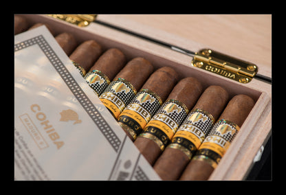 An open wooden cigar box containing several brown cigars with gold and black bands, alongside a branded paper insert. The box is shown in a close-up shot with a visible gold clasp engraved with the brand name.
