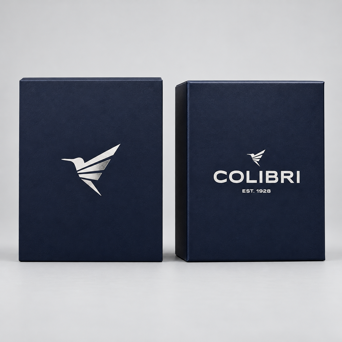 Two dark navy blue rectangular boxes standing upright on a light gray studio background. The left box shows a white bird logo, and the right box shows the white COLIBRI logo with 'EST.