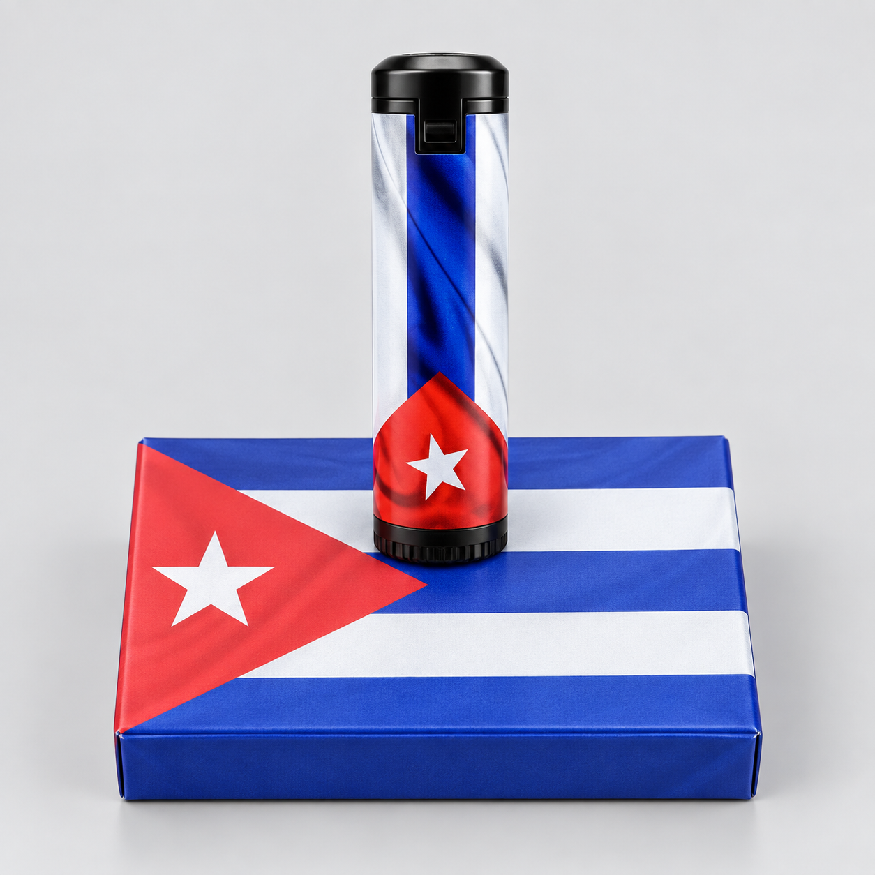 A cylindrical lighter with a Cuban flag design stands upright on top of a Cuban flag-patterned box against a light gray studio background.