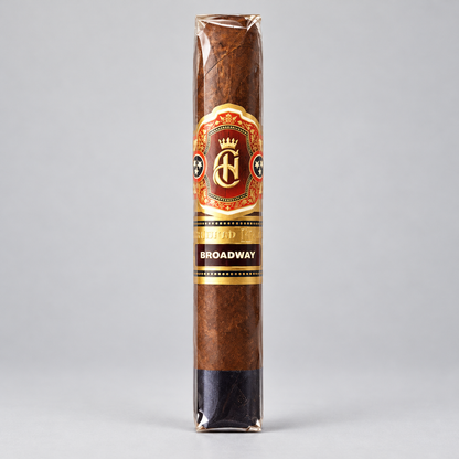 A single wrapped cigar standing upright on a light gray background. The cigar has a brown textured wrapper with a decorative paper label featuring a gold and red crest and the text 'AF' and 'BROADWAY'.
