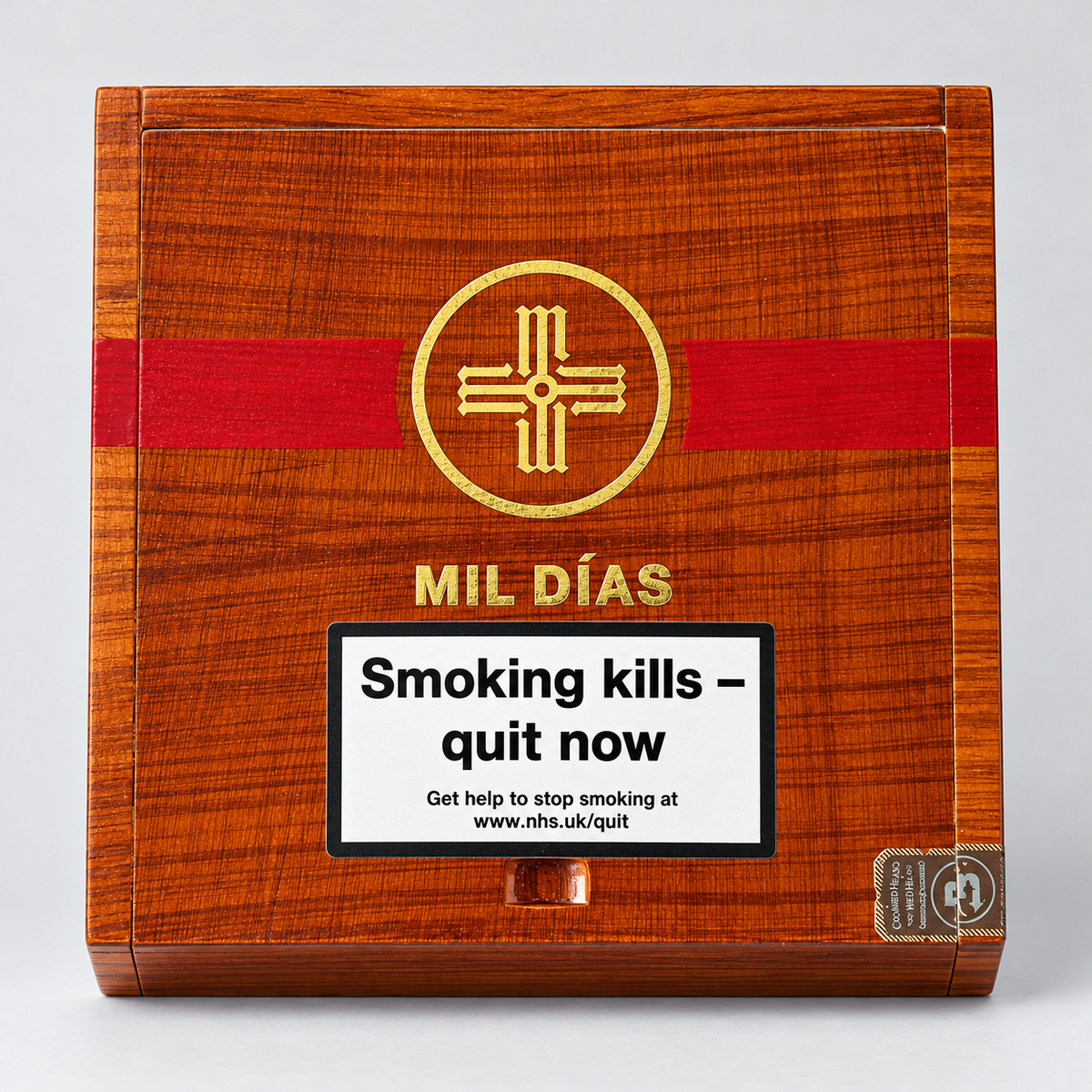 A rectangular wooden box with a warm reddish-brown woodgrain finish, a bold red horizontal band, and a gold circular cross-like emblem. The front also shows the text 'MIL DÍAS' and a large white warning label reading 'Smoking kills - quit now' with smaller quit-smoking contact text.