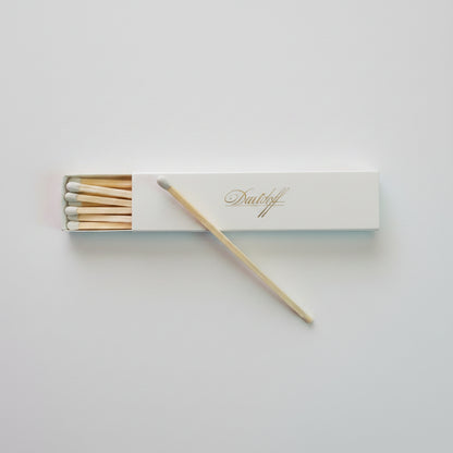 A matchbox with several wooden matches inside, placed on a textured gray surface. A single match rests diagonally across the white box, which has gold cursive branding that appears to read 'D.