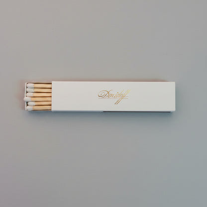 A pack of wooden matches in a white rectangular box with gold foil script, placed on a textured gray surface.