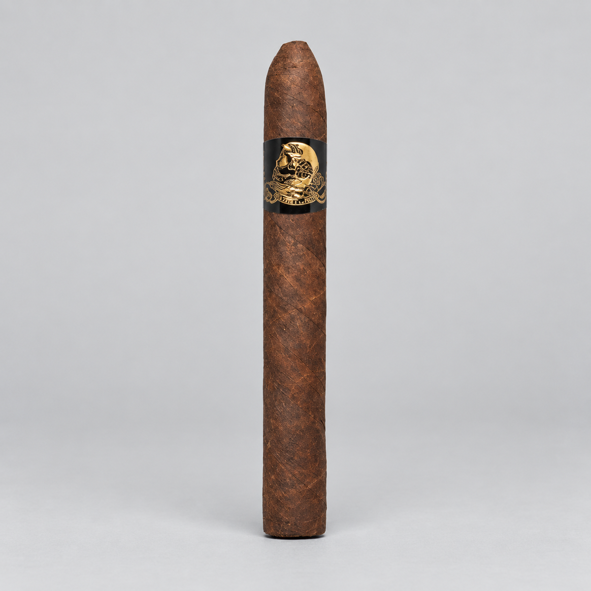 A single brown cigar standing upright against a light gray studio background, featuring a black band with a gold skull-and-crossbones design.