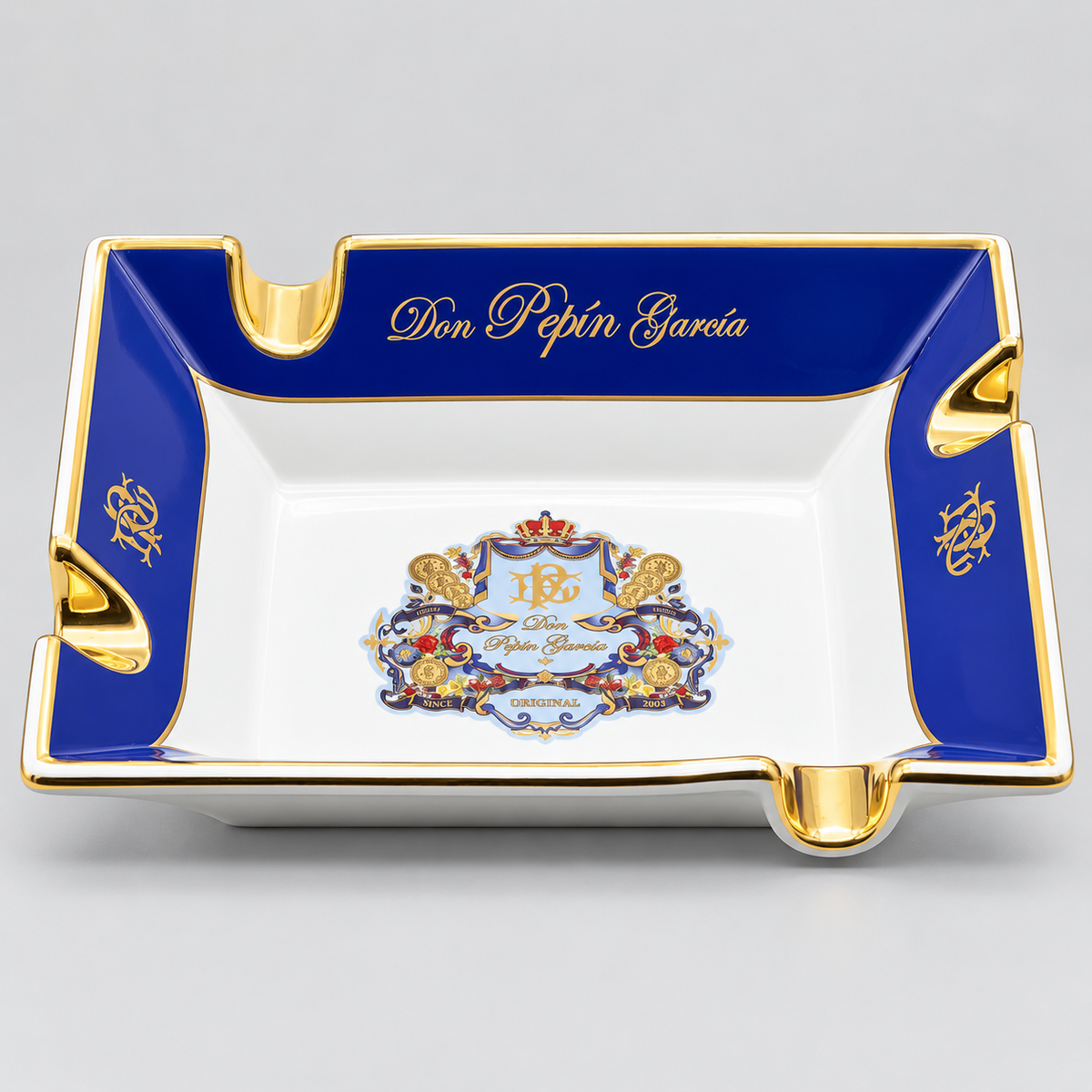 A square white porcelain ashtray with a royal blue border and gold trim, personalized with the name 'Don Pepín García' in gold script. The center features a detailed crest with a red crown, gold lions, and floral elements, and the initials 'PG'.