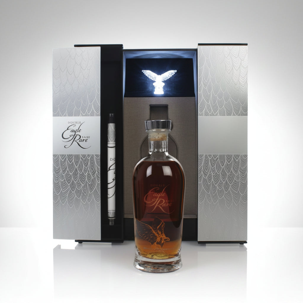 A bottle of amber liquor with a clear glass neck and stopper stands in front of an open luxury presentation box. The box has silver-gray patterned panels, a black center insert, and a small metallic eagle figurine on top.