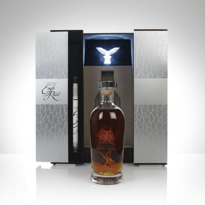 A bottle of amber liquor with a clear glass neck and stopper stands in front of an open luxury presentation box. The box has silver-gray patterned panels, a black center insert, and a small metallic eagle figurine on top.