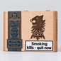 A wooden cigar box with a natural light wood finish, featuring a dark brown engraved lion head emblem, a dark blue vintage-style label on the left, and a white warning label that reads 'Smoking kills - quit now'.
