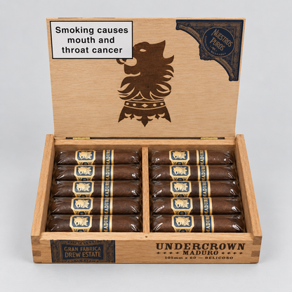 An open wooden cigar box labeled Undercrown Maduro, containing two rows of dark brown cigars wrapped in bands. The inside lid has a large dark emblem and a smoking-health warning label.