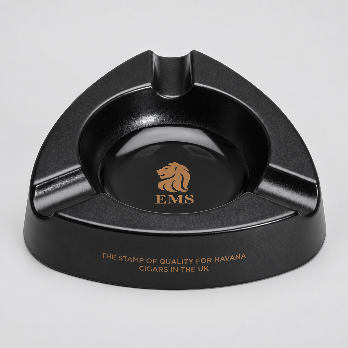 A black triangular ashtray with a glossy finish, featuring a gold lion logo and the letters 'EMS' in the center, and gold text on the front rim reading 'THE STAMP OF QUALITY FOR HAVANA CIGARS IN THE UK'.