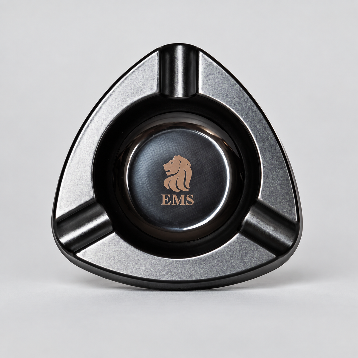 A triangular metal cigar ashtray with a brushed silver and black finish, featuring a central circular insert with a gold lion logo and the letters 'EMS'.
