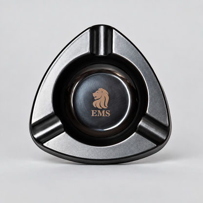 A triangular metal cigar ashtray with a brushed silver and black finish, featuring a central circular insert with a gold lion logo and the letters 'EMS'.